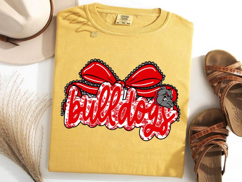 May include: Mustard yellow t-shirt featuring a red bow and the word "bulldogs" in red script. The word "bulldogs" has a floral pattern and a bulldog graphic. A hat and sandals are next to the shirt.