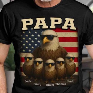 Op de afbeelding: Zwart T-shirt met een afbeelding van een Amerikaanse zeearend met een zonnebril en het woord "PAPA" erboven. Daaronder staan vijf kleinere vogels, ook met zonnebrillen, voor een Amerikaanse vlag.