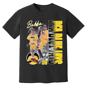 May include: Black t-shirt featuring a baseball player image with the text "Bukka" in yellow script. The shirt also displays "Pea Blue Jays" in vertical yellow letters and the number "38". The design includes a lightning bolt graphic.