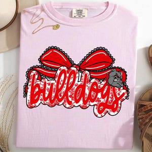 May include: A pale pink t-shirt featuring the word "bulldogs" in red script, embellished with a red bow and a bulldog graphic. The design incorporates a floral pattern. The shirt is folded and displayed with sandals and a hat.