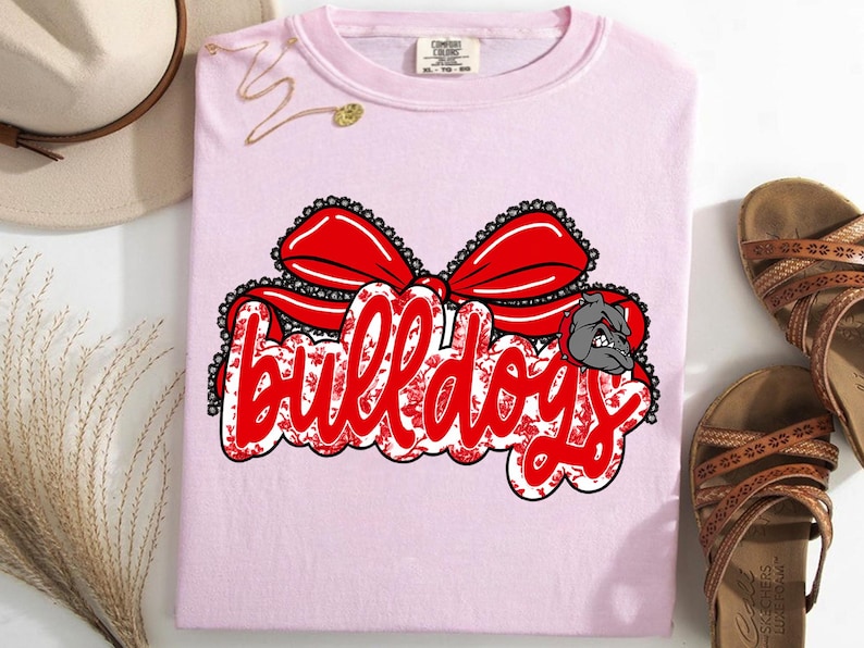 May include: A pale pink t-shirt featuring the word "bulldogs" in red script, embellished with a red bow and a bulldog graphic. The design incorporates a floral pattern. The shirt is folded and displayed with sandals and a hat.