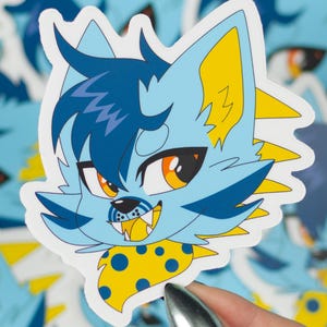 May include: A blue and yellow furry cartoon character with a spotted yellow bandana. The character has a mischievous expression with a toothy grin and bright orange eyes.