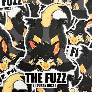 Op de afbeelding: Een verzameling stickers met een gestileerd zwart en geel harig personage in een speelse pose. De stickers hebben de tekst "THE FUZZ" en "(FURRY HUZZ)" in witte letters. Het personage heeft roze en oranje accenten.