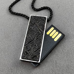 May include: A black USB drive pendant necklace with a textured rectangular pendant and a black chain. The pendant has a silver border and a detailed, geometric design. The USB drive is black with gold-colored contacts.