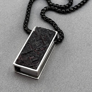 May include: A rectangular pendant necklace with a dark, patterned stone set in a silver frame. The pendant is suspended from a black chain. The stone has a dark red and black color scheme.