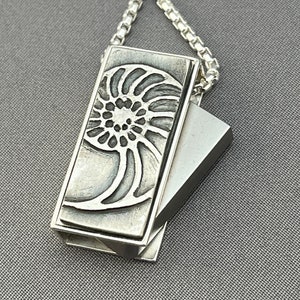 May include: A silver necklace with a pendant featuring a detailed nautilus shell design. The pendant is made of two pieces that slide together.