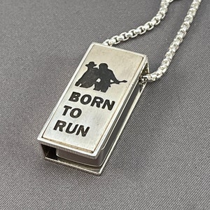 May include: A silver necklace with a rectangular pendant that says "Born to Run" and features a silhouette of two people playing guitars.