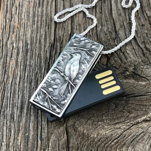 May include: A silver necklace with a rectangular pendant featuring a detailed engraving of a bird on a branch. The pendant is attached to a black USB drive.