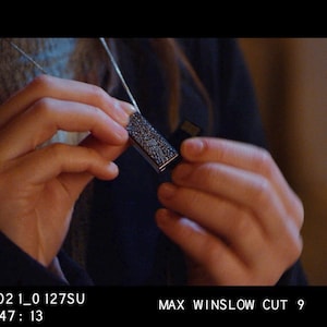 May include: A silver necklace with a rectangular pendant covered in small, black stones. The pendant is being held by a person's hand.
