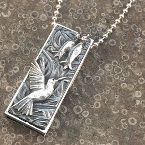 May include: A silver pendant necklace featuring a hummingbird in flight with two fish in its beak. The pendant is rectangular and has a textured surface. The necklace is on a silver chain.