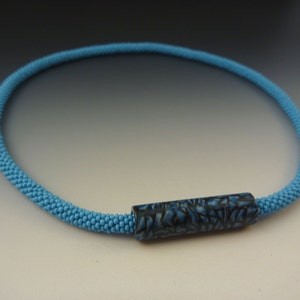 Puede incluir: Un collar de cuentas azul claro con una cuenta cil&iacute;ndrica de patr&oacute;n azul oscuro.