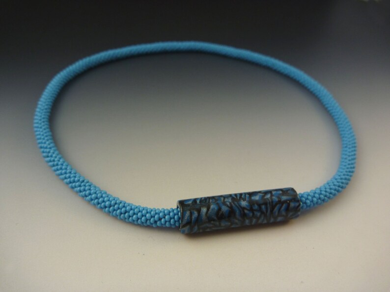 Puede incluir: Un collar de cuentas azul claro con una cuenta cil&iacute;ndrica de patr&oacute;n azul oscuro.