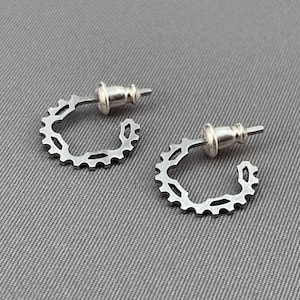 Può includere: Un paio di orecchini a cerchio argentati con un design a forma di ingranaggio. Gli orecchini sono in metallo e hanno un retro a perno.