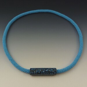 Puede incluir: Un collar de cuentas azul claro con una cuenta cil&iacute;ndrica de patr&oacute;n azul oscuro.
