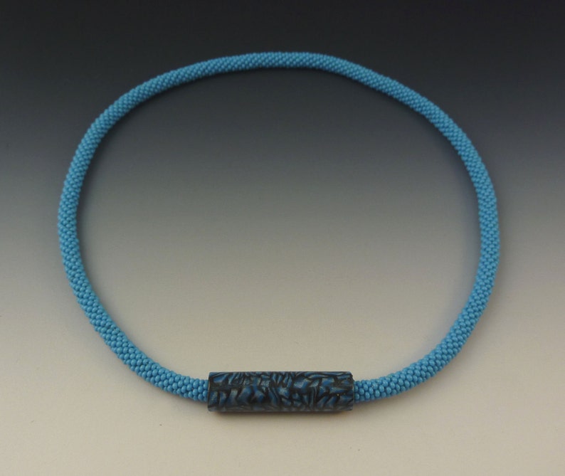 Puede incluir: Un collar de cuentas azul claro con una cuenta cil&iacute;ndrica de patr&oacute;n azul oscuro.