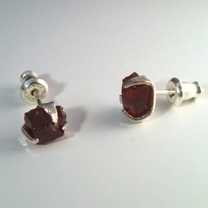 May include: A pair of silver stud earrings with red gemstone accents. The gemstones are rough and have a natural, unpolished look.