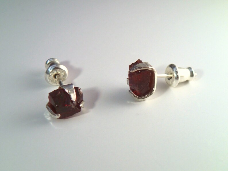 May include: A pair of silver stud earrings with red gemstone accents. The gemstones are rough and have a natural, unpolished look.