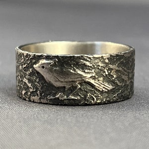 May include: A silver ring with a textured surface and a bird design.