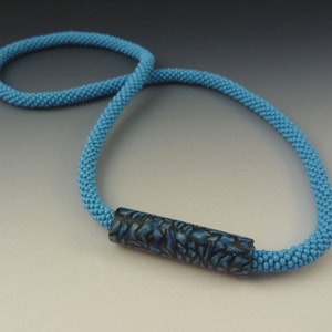 Puede incluir: Un collar de cuentas azul claro con una cuenta cil&iacute;ndrica de patr&oacute;n azul oscuro.