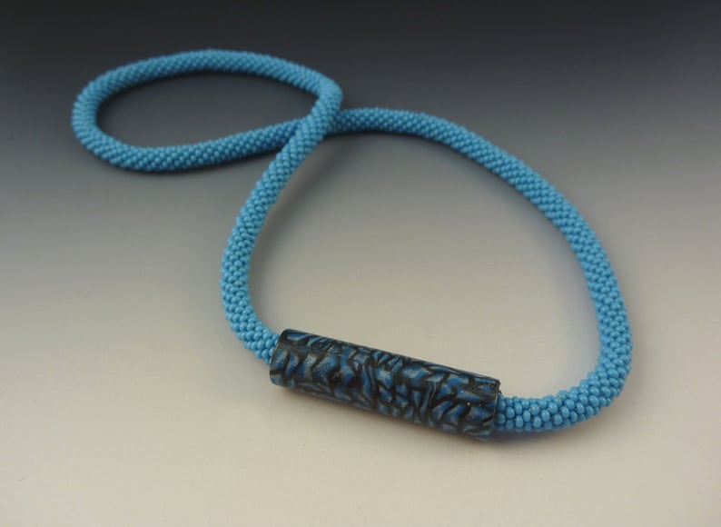 Puede incluir: Un collar de cuentas azul claro con una cuenta cil&iacute;ndrica de patr&oacute;n azul oscuro.