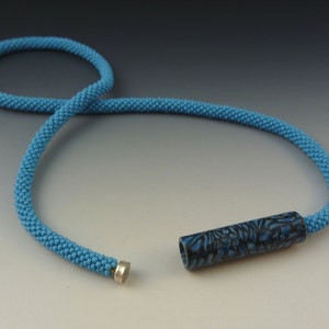Puede incluir: Un collar de cuentas azules con un colgante cil&iacute;ndrico de patr&oacute;n negro y azul. El collar est&aacute; hecho de cuentas peque&ntilde;as, espaciadas uniformemente y tiene un cierre magn&eacute;tico.