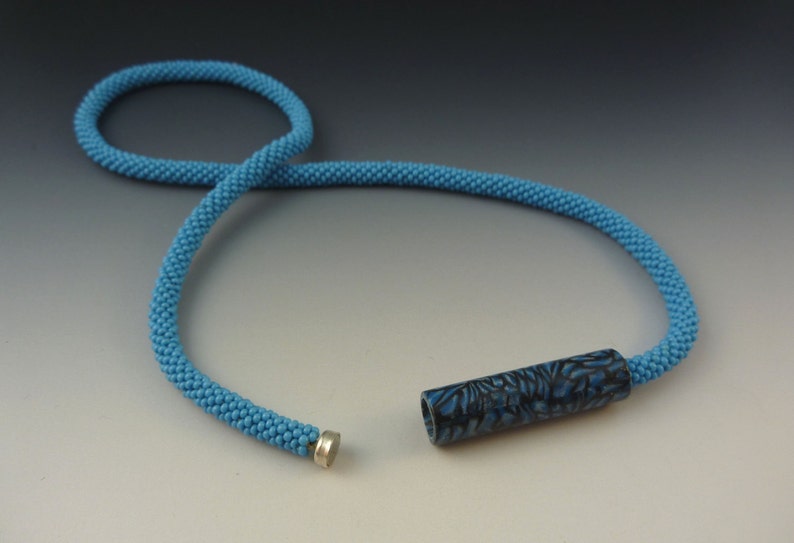 Puede incluir: Un collar de cuentas azules con un colgante cil&iacute;ndrico de patr&oacute;n negro y azul. El collar est&aacute; hecho de cuentas peque&ntilde;as, espaciadas uniformemente y tiene un cierre magn&eacute;tico.
