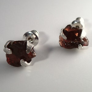 May include: A pair of silver stud earrings with a raw, dark red gemstone. The gemstones are irregular in shape and have a rough, natural texture.