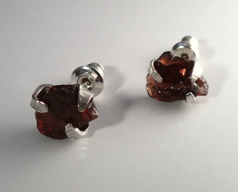 May include: A pair of silver stud earrings with a raw, dark red gemstone. The gemstones are irregular in shape and have a rough, natural texture.