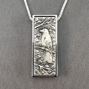 May include: A silver pendant necklace featuring a rectangular pendant with a detailed relief of a bird perched on a branch amidst leaves. The bird is facing left. The pendant is suspended from a delicate silver chain.