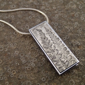 May include: A silver necklace with a rectangular pendant featuring a detailed embossed fern design.