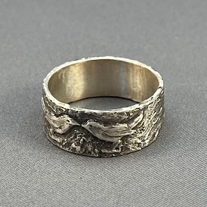 May include: A silver band ring with a textured surface and two birds in flight design.