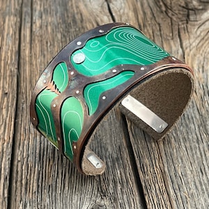 May include: A green and copper cuff bracelet featuring a topographic map design. The bracelet is made of metal and has a leather backing.