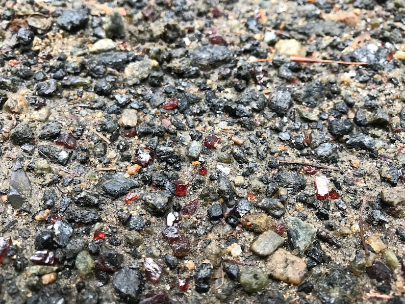 May include: Close-up of a gravel path with small, dark-colored stones and scattered red, shiny pieces of glass.