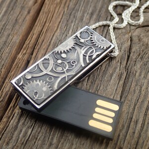 May include: A silver USB flash drive pendant with a detailed gear and swirl design, resting on a wooden surface. The black USB drive is partially open, revealing the gold-colored connectors. A silver chain is attached to the pendant.
