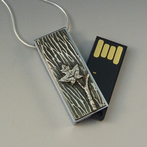 May include: A silver pendant with a textured, bark-like design and a silver maple leaf and branch. The pendant is rectangular and attached to a silver chain. A black USB drive is next to the pendant.