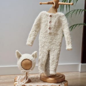 May include: A white knitted baby outfit with a matching hat, displayed on a wooden stand. The outfit has a button closure and the hat has ear flaps.