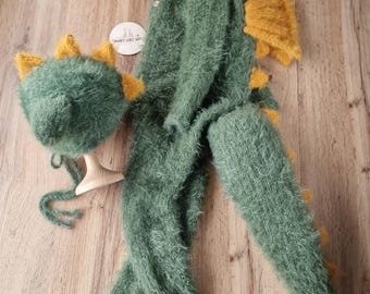 May include: A green and yellow dinosaur-themed baby outfit set. The set includes a hat with spikes, a long-sleeved jumpsuit, and a tail. The outfit is made of a soft, fuzzy material.