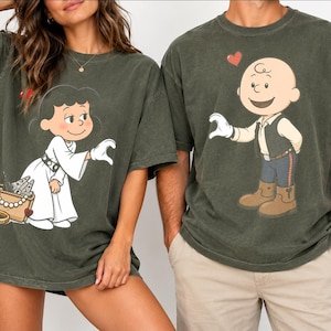 Puede incluir: Dos camisetas de color verde oliva oscuro con personajes de dibujos animados. La camiseta izquierda muestra un personaje con vestido blanco y bolso, la derecha un personaje calvo con chaleco y botas. Ambas tienen pequeños corazones rojos.