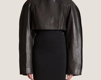 Puede incluir: Bolero de cuero negro con cuello alto y cierre de botón. La chaqueta tiene mangas largas y anchas y un largo recortado, mostrando un diseño moderno. La prenda se lleva sobre un vestido negro.