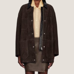 May include: A dark brown suede jacket with a black leather collar and zipper closure. The jacket has four front pockets and silver snap closures. The model is wearing a light yellow button-down shirt and a brown leather skirt.