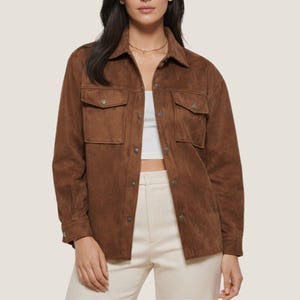 May include: A brown suede-like jacket with two chest pockets and button closures. The jacket has a collared neckline and long sleeves. The model is wearing a white top and cream-colored pants. The jacket is a versatile piece for any wardrobe.