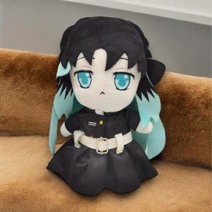 May include: A plush doll of a character with black hair, teal accents, and large blue eyes. The doll wears a black outfit with a white belt and a pleated skirt. The doll is sitting on a brown surface.