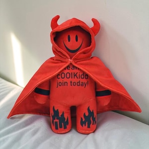 Può includere: Bambola diabolica in peluche rosso con un volto sorridente, corna e un mantello con cappuccio. Il mantello ha il testo "team COOLKids join today!" e fiamme sono stampate sulle gambe della bambola.