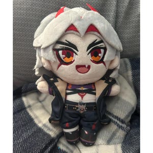 Peut inclure: Poupée en peluche d'un personnage aux cheveux blancs, aux cornes rouges et aux yeux rouges. La poupée porte une veste noire, un haut violet et un pantalon bleu foncé avec une ceinture. La poupée a une expression souriante et est en tissu doux.