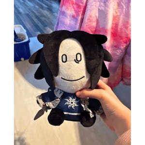 Puede incluir: Muñeco de peluche con cara blanca, pelo negro y un atuendo azul y blanco. El muñeco tiene un diseño sencillo con dos ojos negros y una pequeña sonrisa. El atuendo del muñeco presenta un diseño en forma de sol en el pecho.