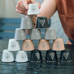 May include: A stack of small metal cups in shades of white, grey, black, and peach. Each cup has a silver rim and a printed design. Some cups display the text "WILD FOREST" and a bear paw symbol.