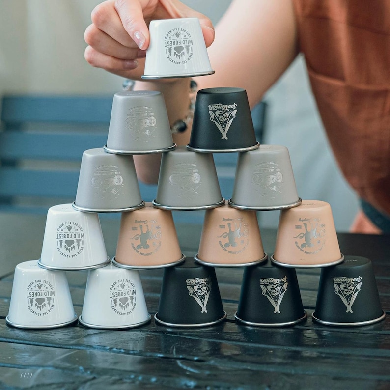May include: A stack of small metal cups in shades of white, grey, black, and peach. Each cup has a silver rim and a printed design. Some cups display the text "WILD FOREST" and a bear paw symbol.