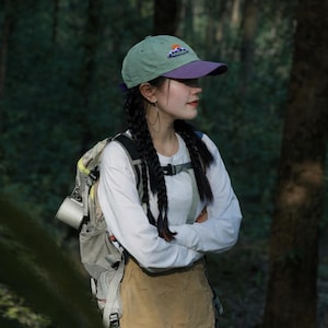 May include: A green and purple baseball cap featuring a mountain and sun design. The cap is worn by a person with long dark braids, a white long-sleeve shirt, and a backpack. The background is a forest.