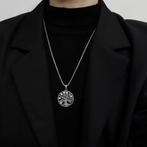 May include: Silver necklace with a circular pendant depicting a tree of life. The pendant has an intricate, open design with small, sparkling details. The necklace is worn over a black turtleneck and a black blazer.