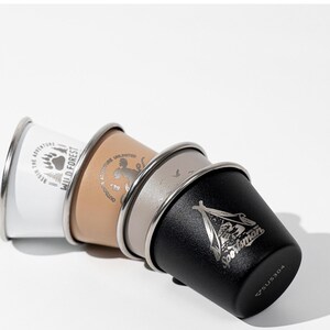 May include: Four stainless steel camping cups in white, tan, grey, and black, stacked together. Each cup has a silver rim and a different outdoor-themed graphic. The white cup says "Wild Forest" and the black cup says "Sportsman".